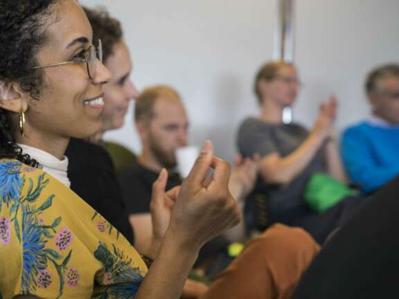 A woman in a yellow floral shirt smiles and claps, with other people blurred in the background, also applauding in a group setting.