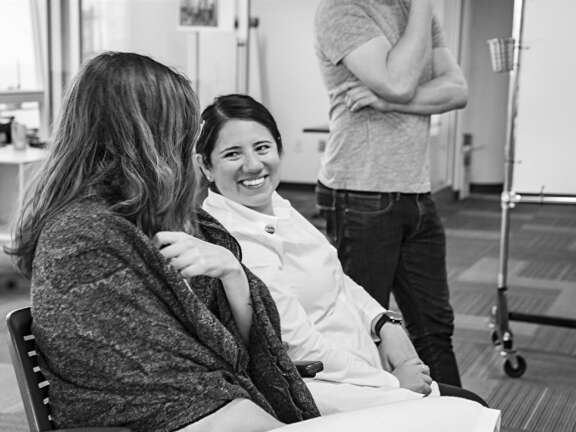 Two women sit and talk, one smiling at the camera, while a man stands nearby with his hand covering his mouth; the setting appears to be a casual indoor office or meeting space. Black and white image.