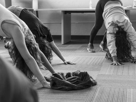 Three people are practicing yoga indoors, positioned in downward dog pose on a carpeted floor. A jacket or sweater lies crumpled on the ground in the foreground. The image is in black and white.