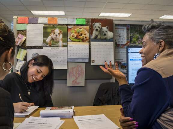 Three women sit at a desk with paperwork; one writes while smiling, another woman gestures as she speaks. Behind them, calendars and animal photos decorate a bulletin board. A computer monitor is visible on the right.
