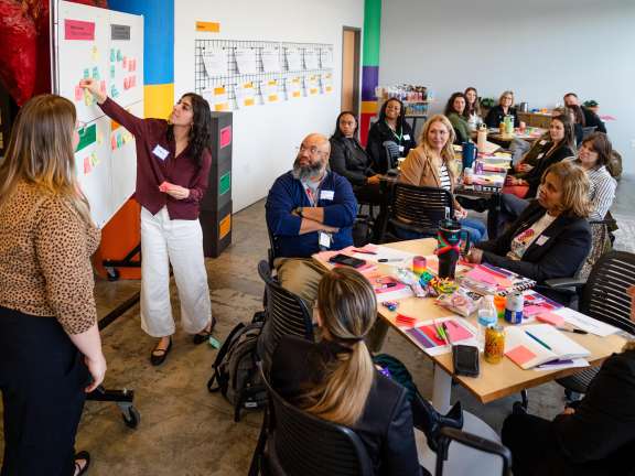 A group of adults sit around tables in a bright meeting room, listening to a woman presenting at a whiteboard covered with colorful sticky notes, as another woman stands nearby. Supplies and papers are spread on the tables.