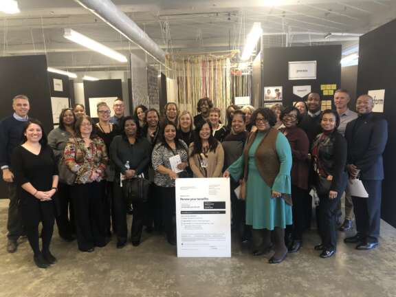 A diverse group of about 30 people stands together indoors, smiling for a group photo. They are gathered around a large poster in a modern office setting with black partitions and signs in the background.
