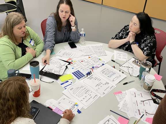 Four women sit around a table covered with papers, sketches, and drinks, collaborating on design documents and brainstorming ideas in a meeting or workshop setting.