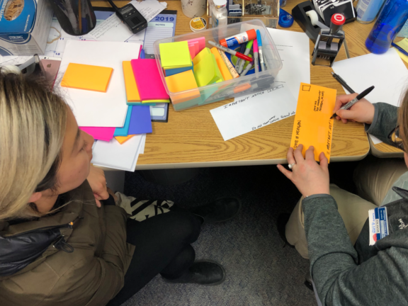 Two people sit at a cluttered desk with colorful sticky notes, markers, and office supplies. One person writes on an orange envelope while the other observes, both engaged in a work-related task.