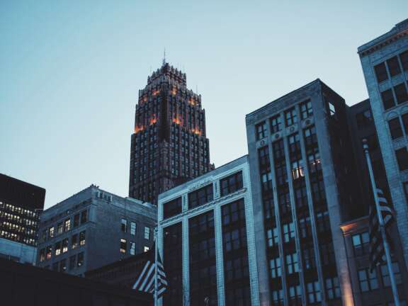 A tall, dark, ornate skyscraper with illuminated windows stands behind shorter, older office buildings against a clear evening sky in a cityscape.