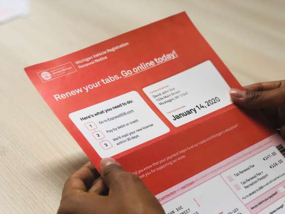 A person holds a red Michigan Vehicle Registration Renewal Notice, detailing steps for renewal, including paying online, by check, or in person. The renewal due date shown is January 14, 2020.
