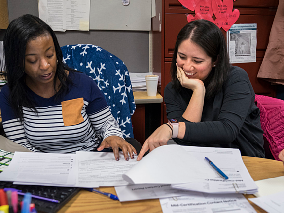 Two women sit at a cluttered desk, reviewing paperwork together. One points at a document while the other smiles, resting her head on her hand. Behind them are bulletin boards and heart-shaped paper decorations.