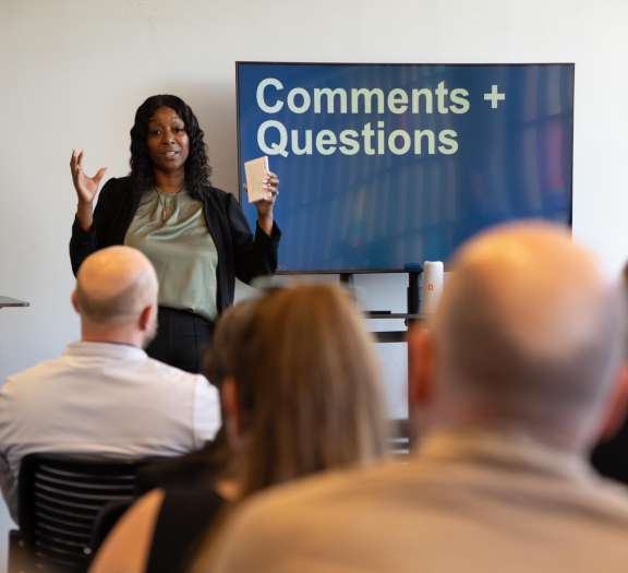 A woman stands in front of an audience, holding a cup and gesturing, with a screen behind her displaying the words Comments + Questions.