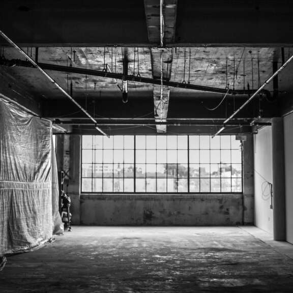 A black-and-white photo of an empty industrial room with a large multi-pane window, exposed ceiling pipes, and a tarp covering part of the left wall. Natural light enters through the window, illuminating the worn floor.