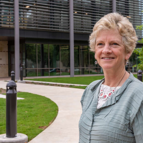 Robin Carnahan with short, light hair and a light green blouse smiles while standing outside a modern building labeled United States Courthouse. The background includes green grass, trees, and a curving walkway.