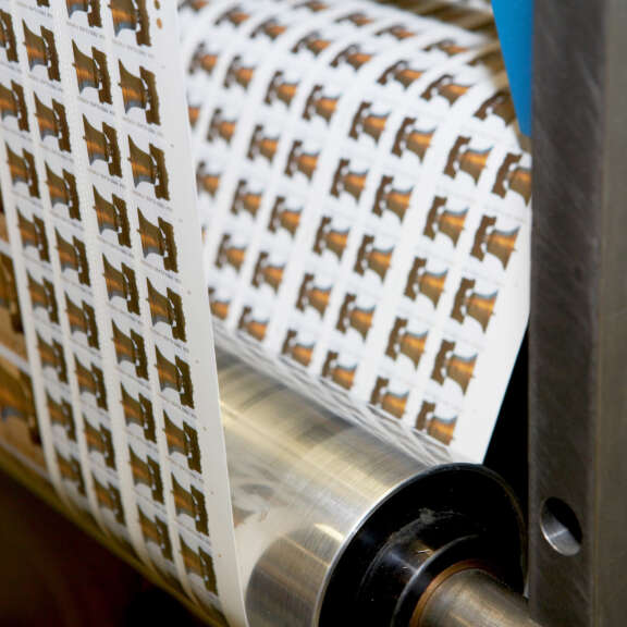 A close-up view of sheets of postage stamps being printed and rolled through a printing press machine. Each stamp features a similar design, and the sheets are aligned in rows.