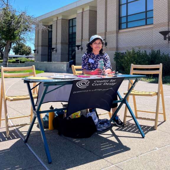A woman sits at a table outside a building with a Center for Civic Design banner. She is wearing a hat and patterned shirt, surrounded by chairs, papers, and supplies on a sunny day.