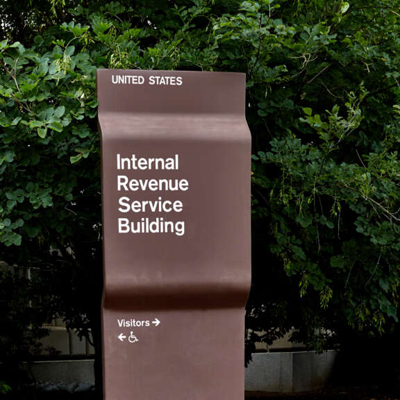 A brown sign with white text reads “United States Internal Revenue Service Building” and points visitors to the right, featuring symbols for accessibility. Lush green trees fill the background.