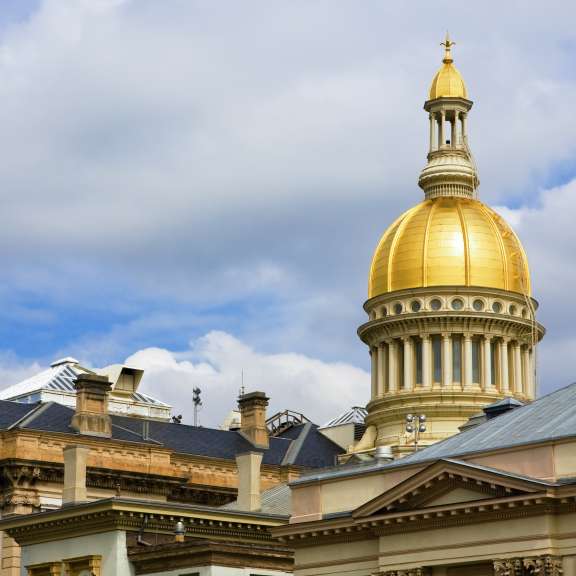 A bright gold dome rises above the historic stone and tan buildings of a government complex, set against a partly cloudy blue sky.