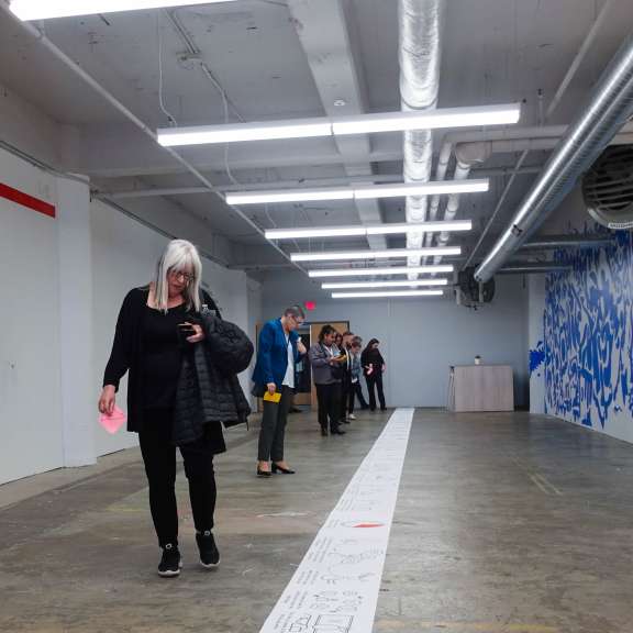 A group of people stands in a line inside a brightly lit industrial gallery space in Civilla's studio, following a journey map, which looks like a long white piece of paper on the floor with drawings; one woman in front looks at her phone while walking.