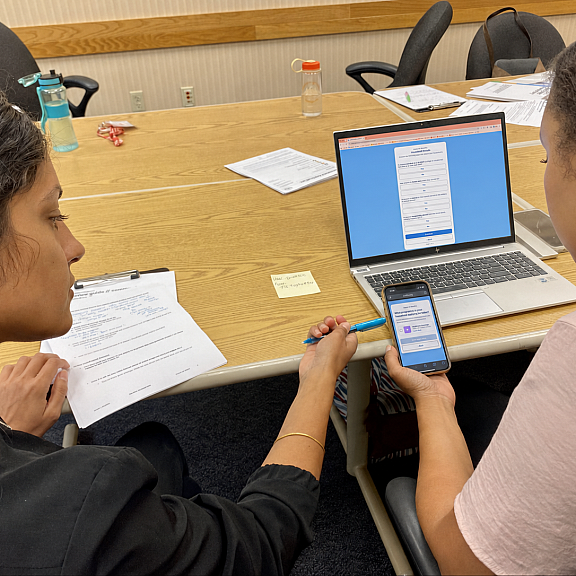 Two people sit at a table with papers, a laptop, and a smartphone. One person points at the phone screen while the other looks on. The laptop displays a form and the workspace has pens and water bottles.