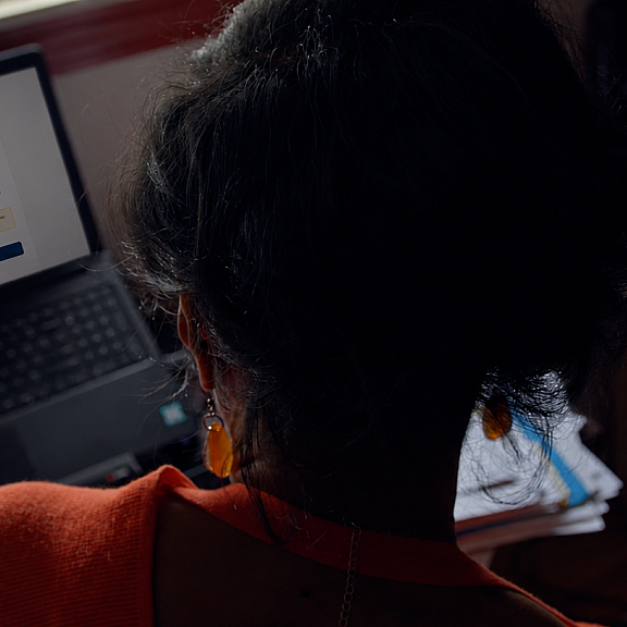A person with dark hair in a bun sits at a desk, looking at a laptop screen displaying a tax or government form. The desk is cluttered with papers, books, and office supplies.