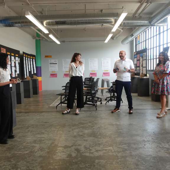 A group of six people stands in a brightly lit room in Civilla's studio  with large windows, listening to a woman presenting next to display boards with images and text. Empty chairs and posters are visible in the background.