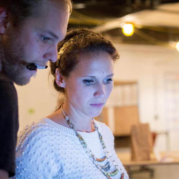 Two people stand closely together indoors, looking intently at a screen. One person wears a headset microphone; the other wears a white sweater and beaded necklace. The background is out of focus with string lights and colorful patterns.