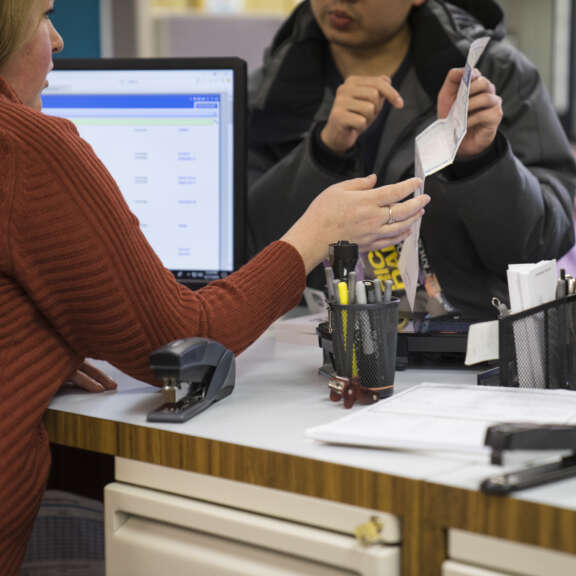 A person sitting at a desk hands paperwork to another person holding ID documents across the counter; a computer monitor, office supplies, and papers are visible on the desk.