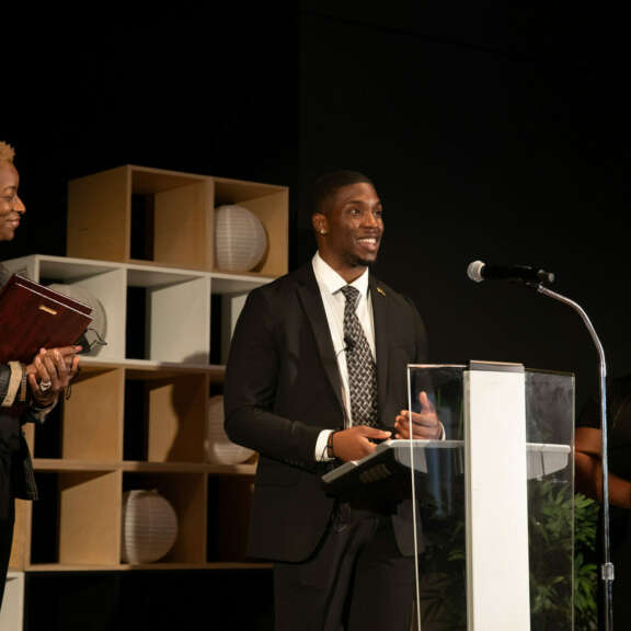 Three people stand on a stage; a man speaks at a podium with a microphone, holding a plaque. Two women beside him, also holding plaques, smile. Shelves with decorative items are in the background.