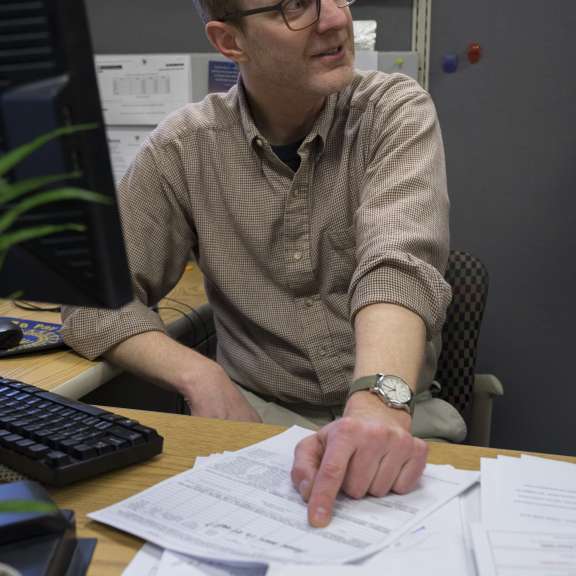 A man wearing glasses and a brown shirt sits at a cluttered desk, pointing at a document. He looks to the side, away from his computer monitor. Papers and a pen are spread out in front of him.