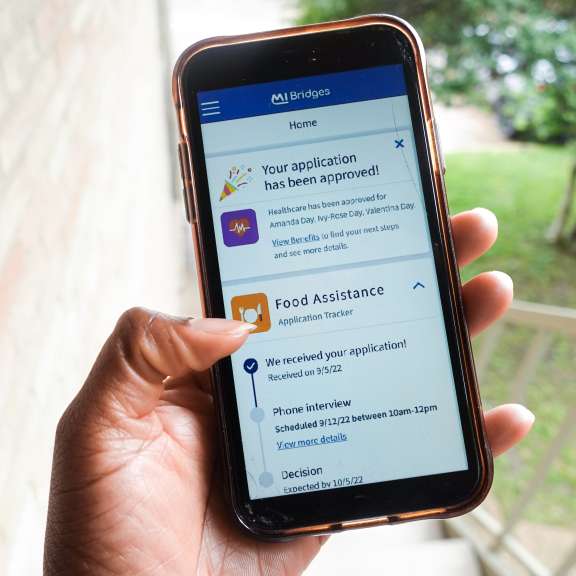 A hand holds a smartphone displaying an approval notification for a health and food assistance application on the MI Bridges app, with stairs and greenery blurred in the background.