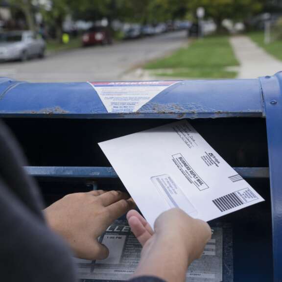 A person’s hand is shown mailing an envelope labeled Official Absentee Ballot into a blue USPS mailbox on a residential street.
