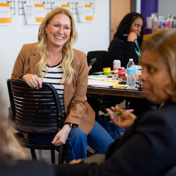 A person with long blonde hair and glasses sits smiling in a classroom during a group activity, surrounded by others engaged in conversation. Papers, drinks, and office supplies are on the table in the background.