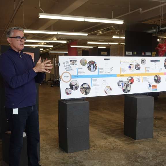 A man wearing glasses and a dark sweater stands indoors, gesturing with his hands while speaking. In front of him is a large timeline display with photos and text, supported by black stands. The room has an industrial feel.