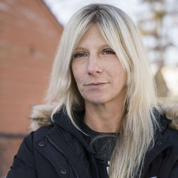 A woman with long blonde hair wears a black winter coat with a fur-lined hood. She stands outdoors in front of a blurred brick wall and bare trees, looking calmly at the camera.