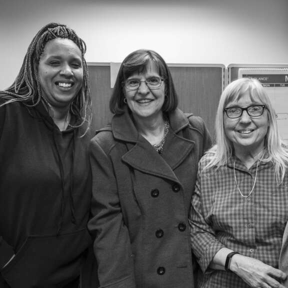 Three women stand close together indoors, smiling at the camera. One wears a hoodie, another a coat and glasses, and the third has long hair and glasses. They appear to be in an office setting. The photo is in black and white.