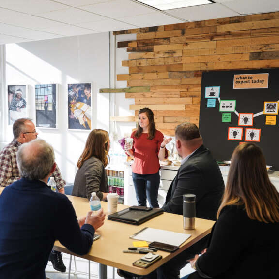 A woman stands by a wall-mounted board, speaking to five seated colleagues in a Civilla studio meeting room. The board behind her displays colorful notes and a “what to expect today” sign. Sunlight streams through nearby windows.