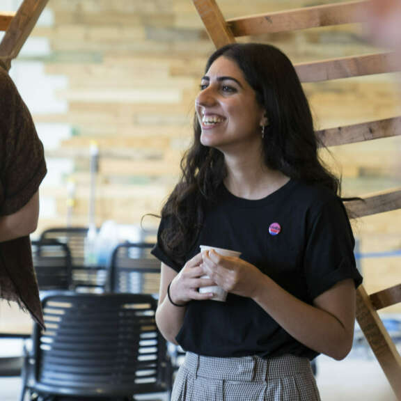 A young woman with long dark hair smiles while holding a paper cup. She wears a black t-shirt and checked pants, standing indoors with wooden slats and chairs in the background.