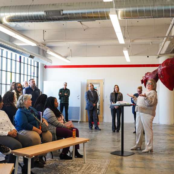 A group of people sit and stand in a brightly lit room, attentively listening to a woman speaking at a podium. Posters are displayed on the wall and a large red sculpture is in the background.