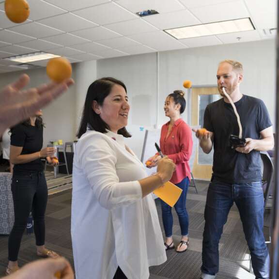 Several people stand in an office space, smiling and interacting. One person juggles oranges, while others hold papers and oranges, appearing to participate in a group activity or team-building event.
