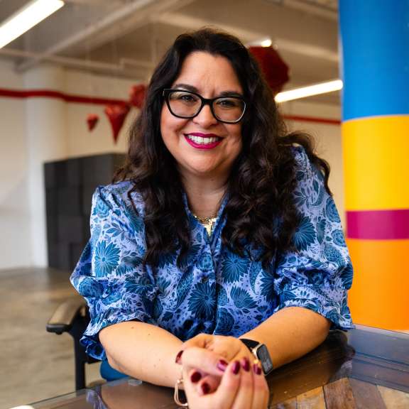 A woman with long dark hair and glasses, wearing a blue floral blouse, sits at a glass table and smiles. The background features a colorful pillar and an open, modern office space.