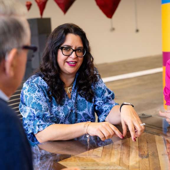 Three people sit around a wooden table having a discussion. The woman in the center gestures while talking, wearing glasses and a blue patterned shirt. Another woman in pink listens attentively; a man with gray hair is seen from behind.