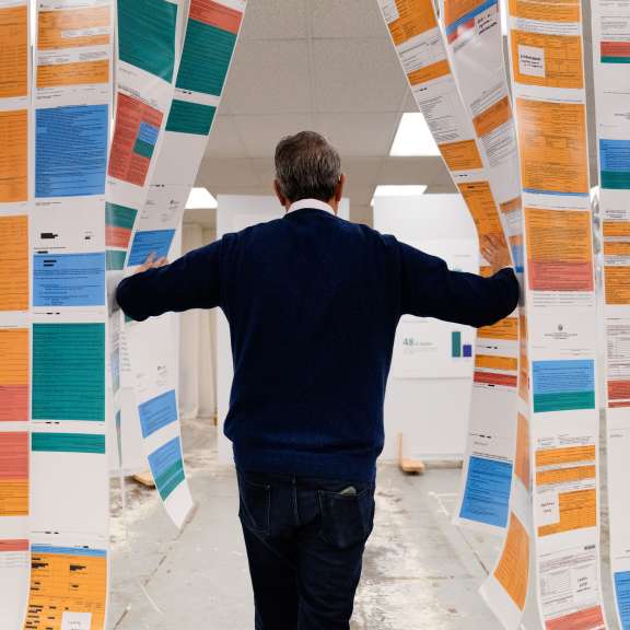 A person in a blue sweater stands with their back to the camera, walking through hanging sheets of colorful printed documents in an indoor setting.