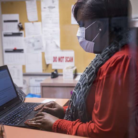 A woman wearing a face mask, red top, and scarf types on a laptop at a reception desk. Office documents and a “DO NOT SCAN” sign are visible on the bulletin board in the background.