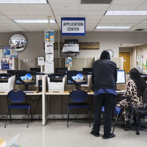Two people use desktop computers at an application center, with several workstations, chairs, bulletin boards, and a sign overhead reading APPLICATION CENTER in a well-lit room.
