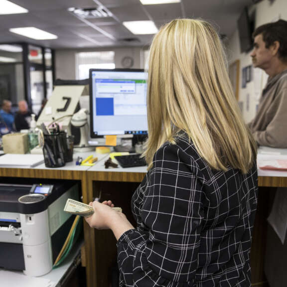 A woman with blonde hair holds cash at a counter in an office setting, facing a computer screen. Another person stands nearby, and several people are visible in the background waiting.