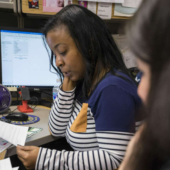 A woman with long dark hair, wearing a striped shirt, sits at an office desk reading papers. A computer monitor, a small fan, and office supplies are on the desk. Another person is partially visible in the foreground.