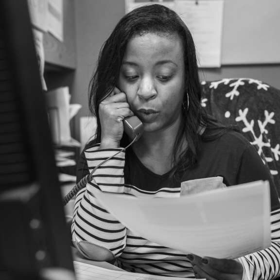 A woman sits at a desk, holding a phone to her ear while reading documents. She appears to be engaged in a work-related call, surrounded by office papers, a computer monitor, and a bulletin board behind her.
