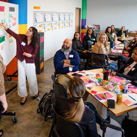 A group of adults sit around tables in a bright meeting room, listening to a woman presenting at a whiteboard covered with colorful sticky notes, as another woman stands nearby. Supplies and papers are spread on the tables.