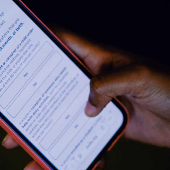 A person holds a smartphone displaying a survey form with selectable answers. The screen shows questions under the household details section. The background is dark, and only the hand and phone are clearly visible.