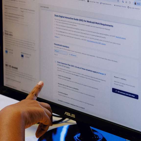 A person points at a computer monitor displaying an informational webpage, with a laptop and colorful cup visible in the background on a desk.