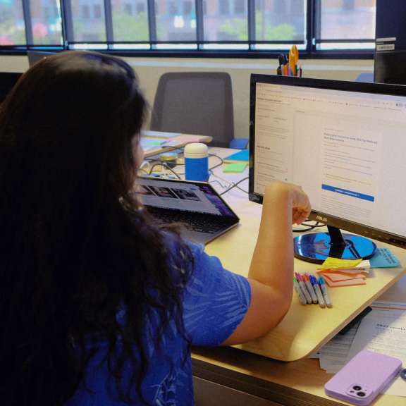 Two people sit at a desk looking at a computer screen together, with notebooks, pens, a laptop, a water bottle, and a phone on the desk in a brightly lit office with large windows.