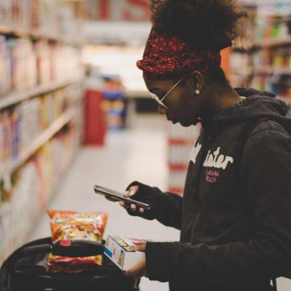A person wearing glasses and a red headband stands in a grocery store aisle, holding a box and using their phone. Their shopping cart contains bags of snacks, and shelves of products line both sides of the aisle.