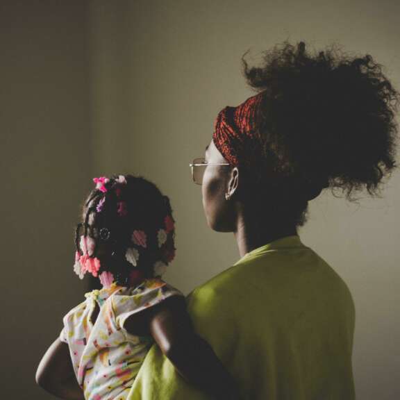 A woman with curly hair in a red headband holds a young girl with beaded braids. Both are seen from behind, looking toward a softly lit window in a dimly lit room.